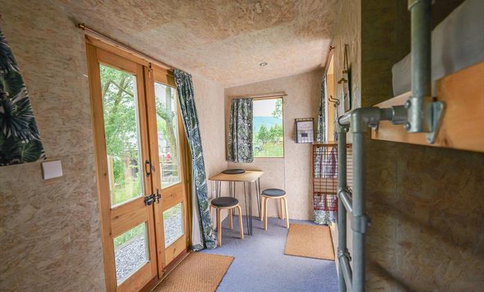 Inside a wooden cabin with double doors and a window, both with curtains. On the floor there is blue carpet, 2 door mats and a small table